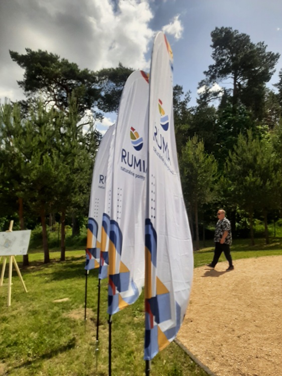 Flagi miasta Rumi z napisem "naturalnie pomysłowa" na tle zielonych drzew podczas konferencji prasowej w parku przy ulicy Polnej