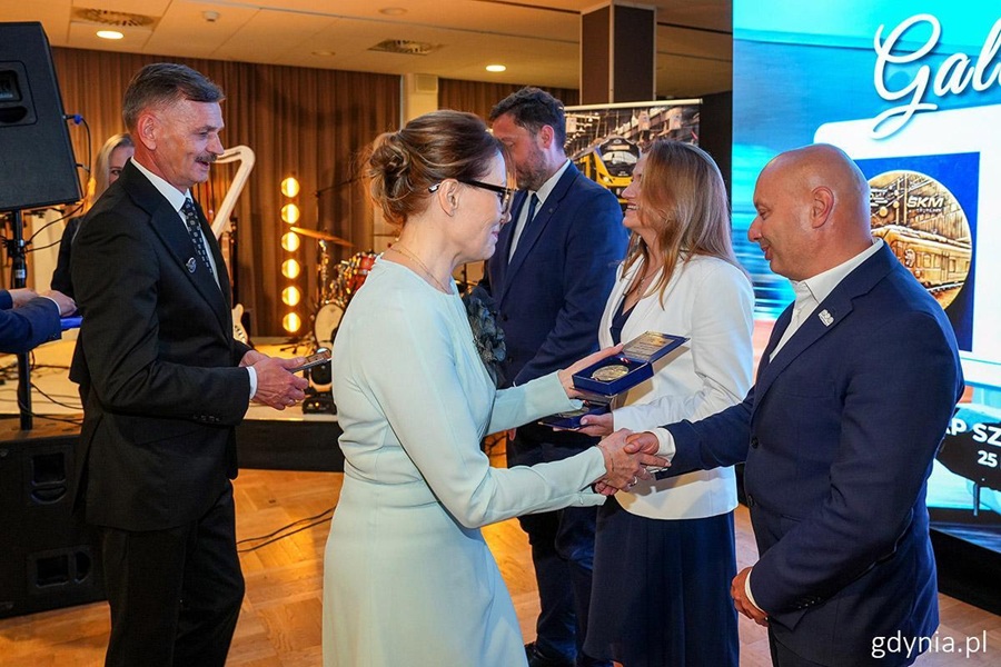 Wręczenie medalu okolicznościowego podczas Gali Jubileuszowej PKP SKM Trójmiasto z okazji 25-lecia spółki. Kobieta w jasnoniebieskim stroju podaje medal mężczyźnie w granatowej marynarce, w tle ekran z logo SKM.