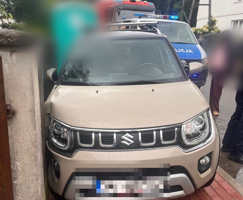 Suzuki Ignis wciśnięte między ogrodzenie posesji a mur po kolizji na ulicy Leśnej w Redzie — w tle radiowóz policji z włączonymi sygnałami świetlnymi